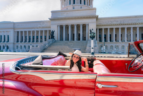 Photography Young Latina woman with sunglasses and a hat smiles while sitting in a vintage red car near El Capitolio in Havana, Cuba