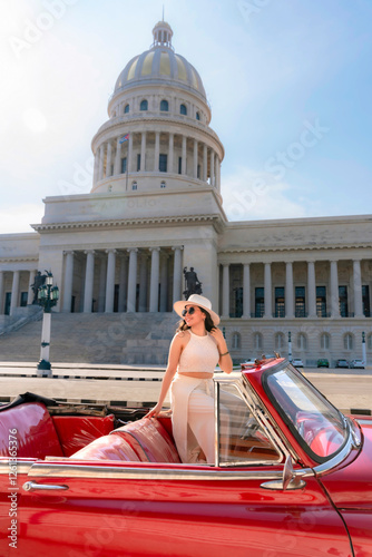 Photography Young Latina woman with sunglasses and a hat smiles while sitting in a vintage red car near El Capitolio in Havana, Cuba