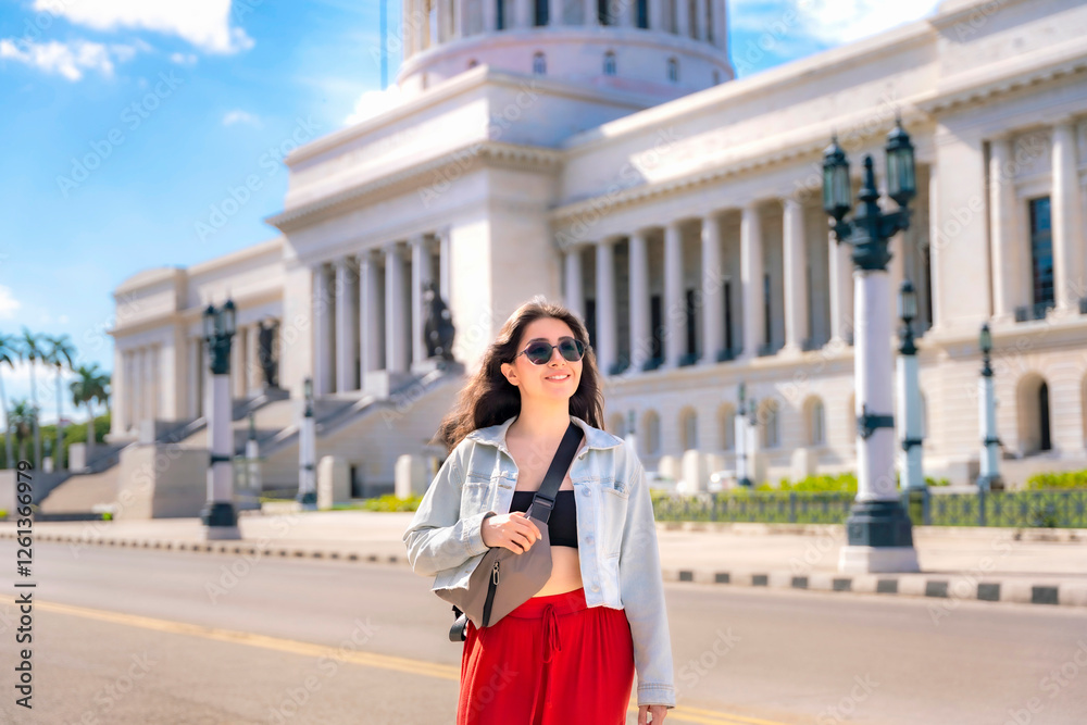 Fototapeta premium A stylish woman in sunglasses and a red skirt walks past El Capitolio in Havana, Cuba, on a sunny day with blue skies. 