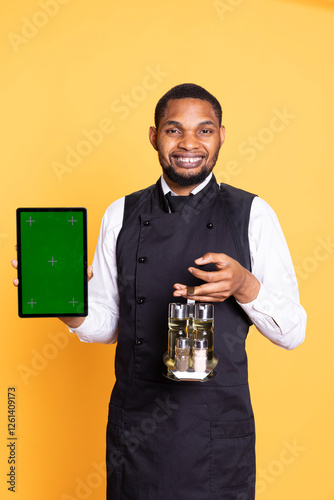 Smiling waiter in uniform presenting a device with isolated chroma key screen, carrying a cruet set to serve clients. Catering industry employee showing mockup display in studio. photo