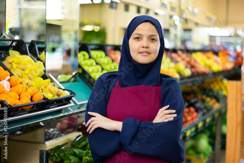 Young woman seller in hijab posing near shelves with vegetables and fruits in grocery store photo