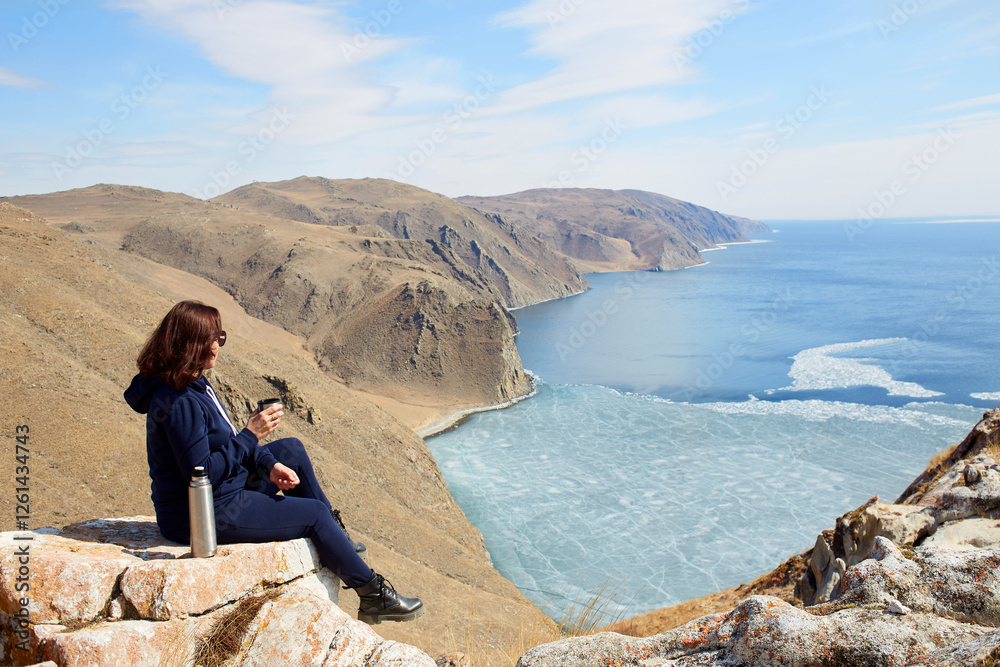 A rest in nature with a beautiful view of the lake in spring. A female traveler is sitting on a rock and drinking hot tea from a thermos. Freedom, travel concept, active recreation, hikking A rest in nature with a beautiful view of the lake in spring. A female traveler is sitting on a rock and drinking hot tea from a thermos. Freedom, travel concept, active recreation, hikking