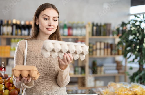 Young beautiful woman shopper in casual clothes chooses package of eggs in big supermarket
