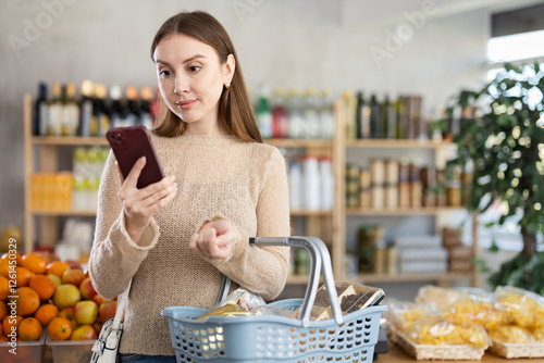 Visiting store, girl scrolling phone screen and looking for something in showcase of shop. She has packed basket of groceries, and is looking at imported delicacies in window photo