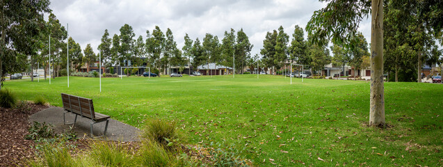 panoramic view of a public park with well-maintained green lawn, bordered by tall eucalyptus trees and residential homes in the background. A wooden bench on side, offering a peaceful resting spot.