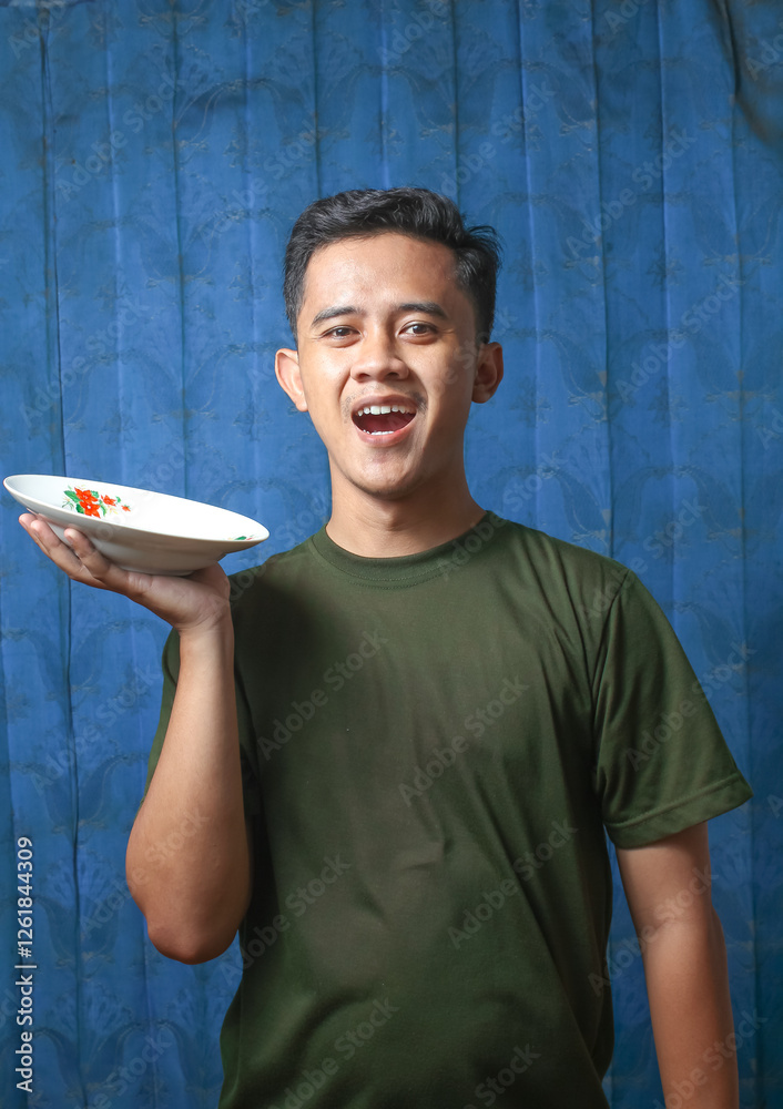 Asian man holding plate with cheerful expression on blue curtain background Asian man holding plate with cheerful expression on blue curtain background