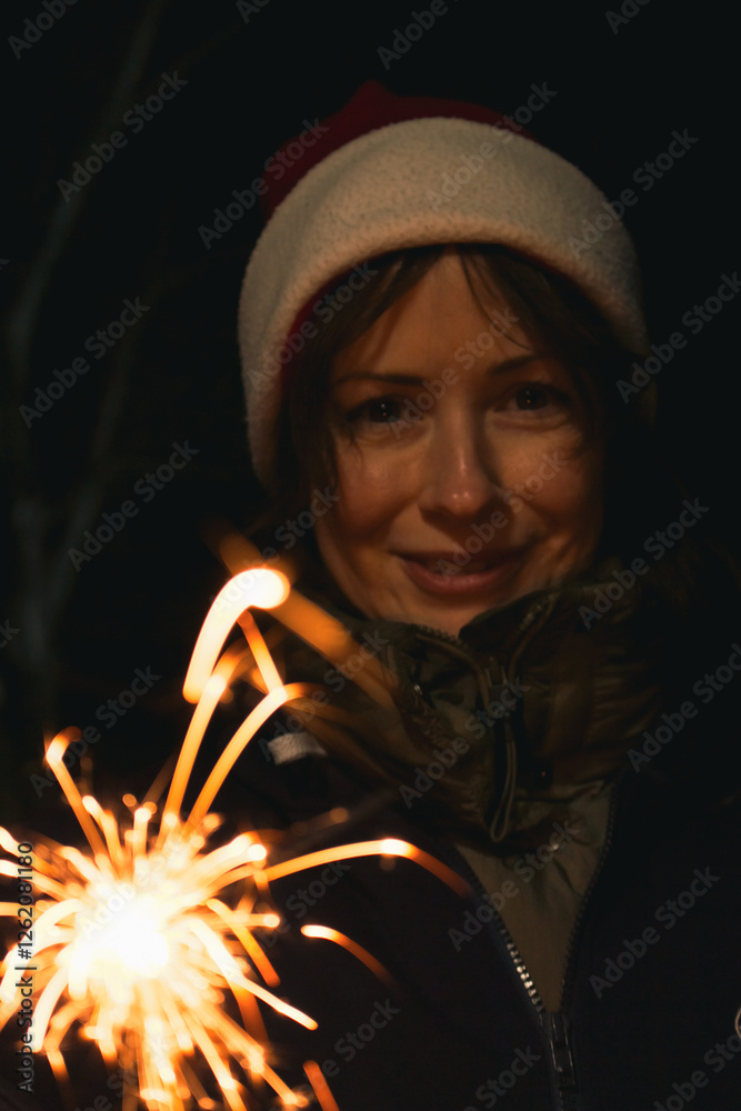 Girl painting with freeze light. Festive freezing lights in the night. Sparkling forms made by woman in Santa hat. Stop light drawings. New Year fun. Firework background. Freeze light symbol in motion