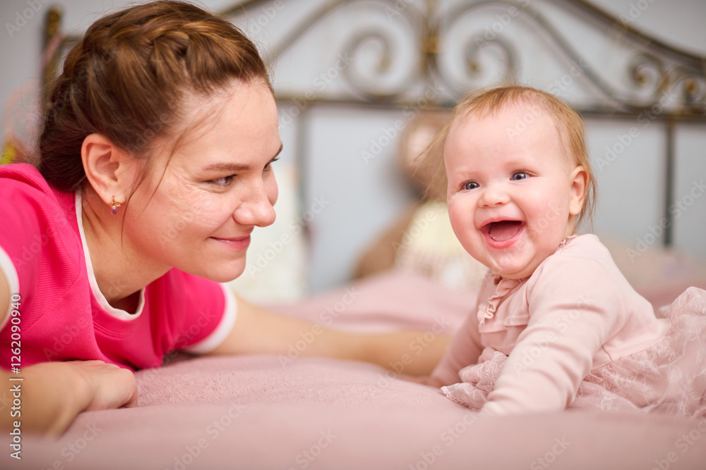 Young woman with brown hair, smiling baby girl indoors. Warm pink tones, soft lighting, metal bedframe in background. Expression of joy and connection Young woman with brown hair, smiling baby girl indoors. Warm pink tones, soft lighting, metal bedframe in background. Expression of joy and connection