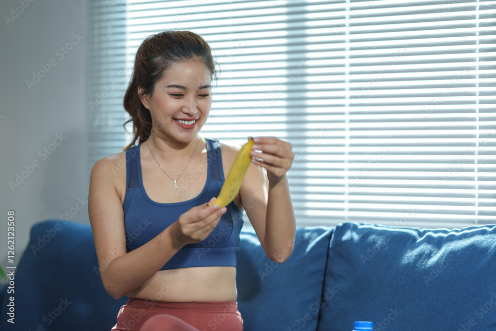 Young woman in activewear holding a banana, sitting on a blue couch. Sunlight filters through blinds, creating a relaxed and healthy lifestyle atmosphere. A water bottle is nearby Young woman in activewear holding a banana, sitting on a blue couch. Sunlight filters through blinds, creating a relaxed and healthy lifestyle atmosphere. A water bottle is nearby