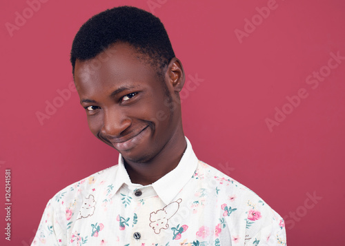 Close-up headshot of a young man in a floral shirt making a weird, awkward smile against a coral background. His playful and humorous expression adds a quirky and lighthearted feel to the portrait.