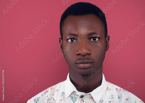 Close-up headshot of a young man with short black hair, wearing a floral shirt, making a neutral expression. His calm and composed face is set against a coral background in a studio portrait.