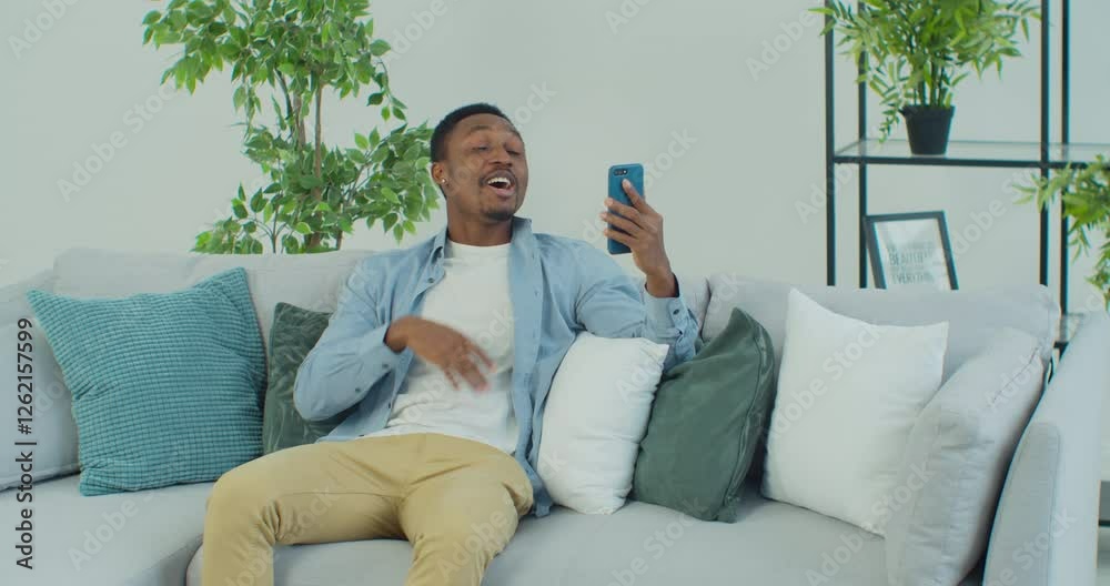 Young African American man with smartphone making video call while sitting on couch in living room. Portrait of happy man mobile video calling , sitting on sofa at home. Young African American man with smartphone making video call while sitting on couch in living room. Portrait of happy man mobile video calling , sitting on sofa at home.