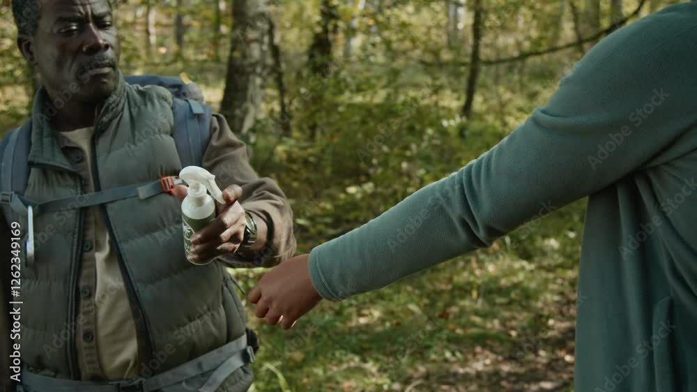 African American father wearing backpack spraying insect repellent on hiking clothes of teenage daughter on forest path African American father wearing backpack spraying insect repellent on hiking clothes of teenage daughter on forest path