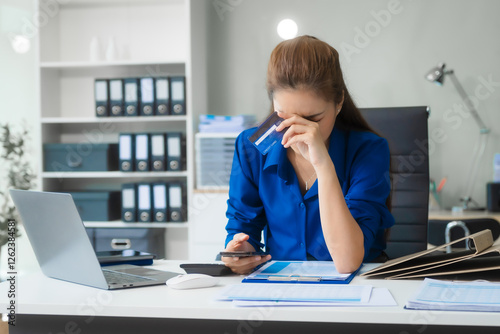 An Asian businesswoman in a blue formal shirt is working at her desk, analyzing financial charts on a laptop and mobile phone, focusing on credit management and financial strategies. photo