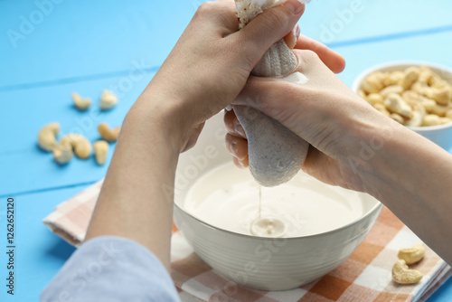 Woman straining cashew milk into bowl at light blue wooden table with nuts, closeup photo