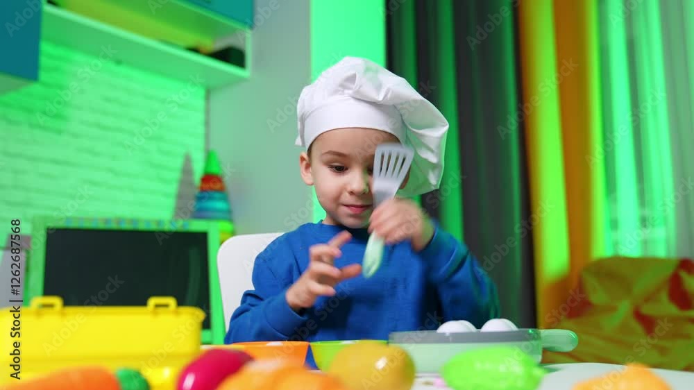 Cute Caucasian boy in chef cap sits at desk holding a plastic kitchen spatula. Baby cook plays with crockery and vegetables at home. Cute Caucasian boy in chef cap sits at desk holding a plastic kitchen spatula. Baby cook plays with crockery and vegetables at home.