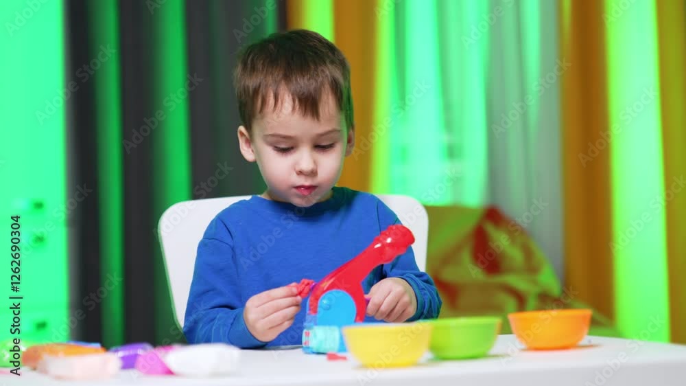 Focused baby boy in blue sweater sits at desk busy with plasticine. Lovely child is busy with his hobby at home. Focused baby boy in blue sweater sits at desk busy with plasticine. Lovely child is busy with his hobby at home.