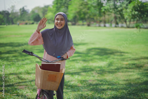 Young Asian beautiful woman standing beside bike at public park aand green grass in morning, looking looking someone and waving hand with smile. photo