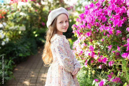 Beautiful caucasian little girl in greenhouse among bush colorful azalea flowers. Portrait of a child girl wearing pink dress and wear beret in the botanical garden arbor among the blossom flowers.	
