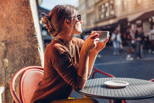 Young stylish woman sits on cafe terrace outdoor drinking coffee. Woman enjoys architecture of European city on street photo