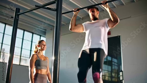 A male athlete with a left knee injury shows determination in pull-ups, guided by a trainer in a brightly lit gymnasium, focusing on improvement, performance and goals.