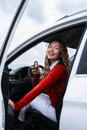 Young smiling overjoyed woman in casual wear holding a key to her new car. Happy female owner of new automobile showing a key and looking at camera. Outside of car view. Buy or rent auto. Vertical.