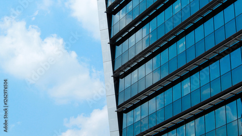 A modern glass building reflects the blue sky and clouds. Its sleek design and geometric patterns create a striking urban aesthetic