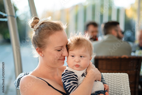 Portrait of happy mother with her handicapped baby in a cafe outside. Young blonde mom holding her son with cerebral palsy. Special needs baby boy. Diversity and inclusion concept.
