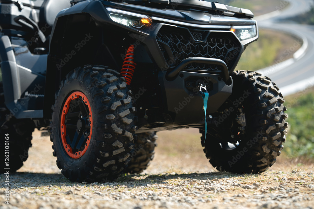 A close-up of a rugged all-terrain vehicle, showcasing its bold tires and sleek design, parked on a gravel path with a winding road in the background. A close-up of a rugged all-terrain vehicle, showcasing its bold tires and sleek design, parked on a gravel path with a winding road in the background.