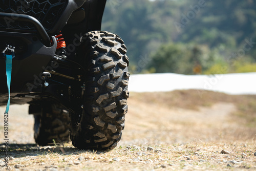A close-up view of an all-terrain vehicle tire on a gravel path, showcasing its rugged tread and suspension detail against a blurred natural background.