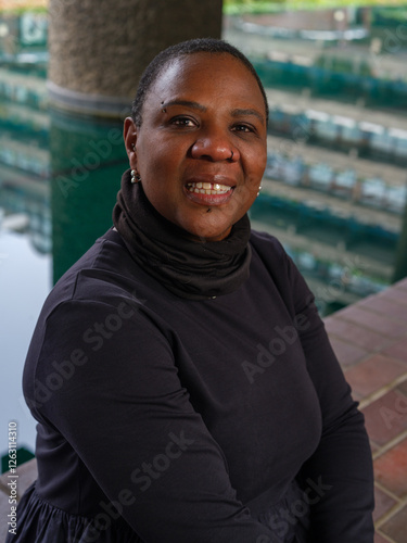 A strong and confident woman gazes directly at the camera, wearing a black outfit with a high collar. The blurred urban background enhances the portrait’s depth