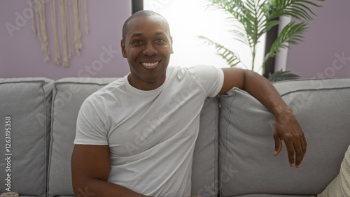 Young man sitting on a couch in a cozy living room, smiling warmly and exuding confidence in a casual home setting enhanced by soft neutral tones and decorative plants.