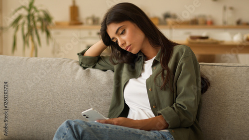 Boring sad upset frustrated stressed Arabian woman Indian girl female Hispanic lady exhausted sorrowful disappointed holding mobile phone smartphone unwanted pregnancy problem thinking at home couch photo