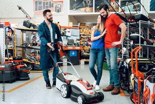 Slika na platnu Salesman in blue robe is showing couple of clients new lawn mover in power tools store