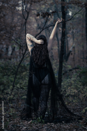 This striking photograph features a woman standing in a dark, moody forest, exuding a gothic and mysterious aura. She is dressed in an all-black ensemble, including a sheer, flowing cape.