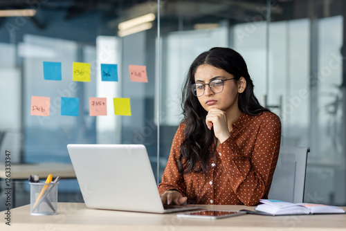 Serious thinking woman at work with laptop inside office. Businesswoman sitting and reading data from screen. Employee thoughtfully solving current problems.