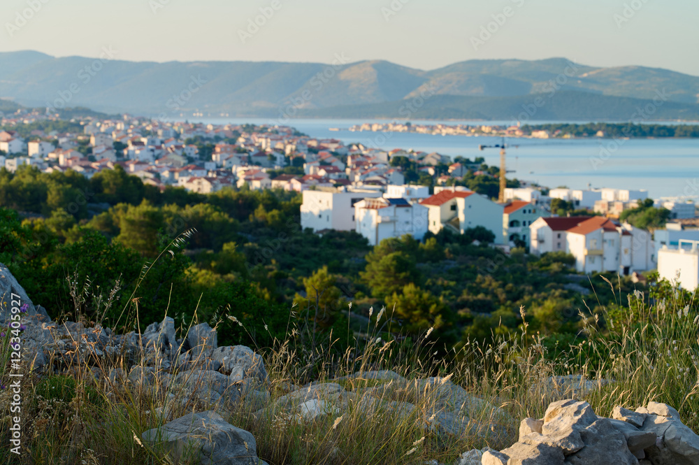 Fototapeta premium Golden sunlight bathes a serene coastal village, nestled among green hills, as the calm sea glimmers. Houses with red roofs create a picturesque landscape filled with tranquility.