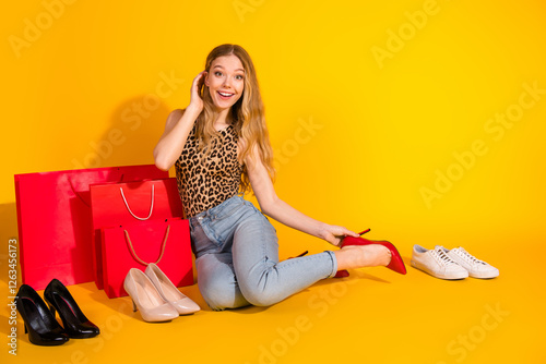 Cheerful young woman in leopard print top sitting with shopping bags and shoes on bright yellow background