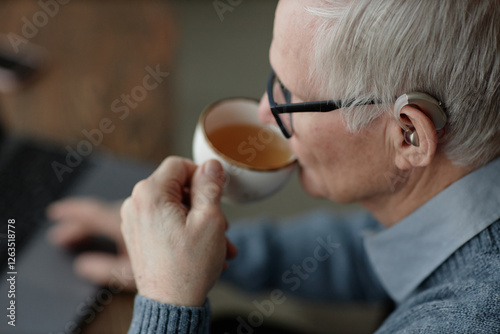 Side view of elderly deafened man wearing hearing aid behind ear using laptop while enjoying warm tea drink at home, copy space photo