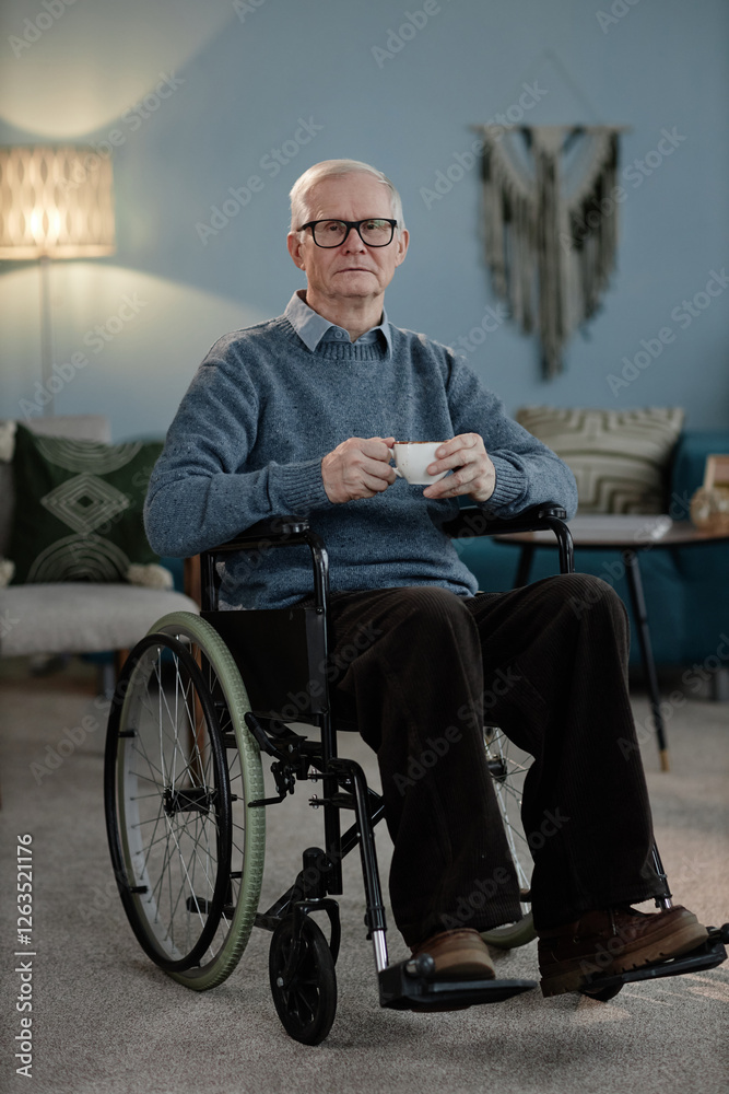 Vertical full length shot of senior male wheelchair user wearing blue sweater looking at camera while holding cup of tea in cozy home setting Vertical full length shot of senior male wheelchair user wearing blue sweater looking at camera while holding cup of tea in cozy home setting