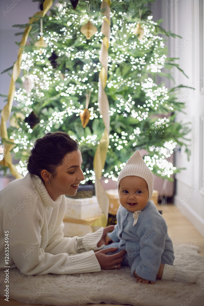 A mother in a white sweater sits with her daughter for 1 year on the floor by the Christmas tree in an apartment on New Year's Eve