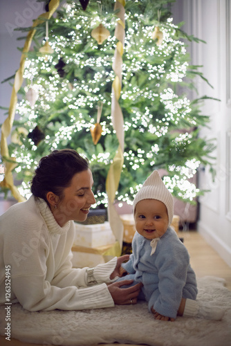 Obraz na płótnie A mother in a white sweater sits with her daughter for 1 year on the floor by the Christmas tree in an apartment on New Year's Eve w salonie