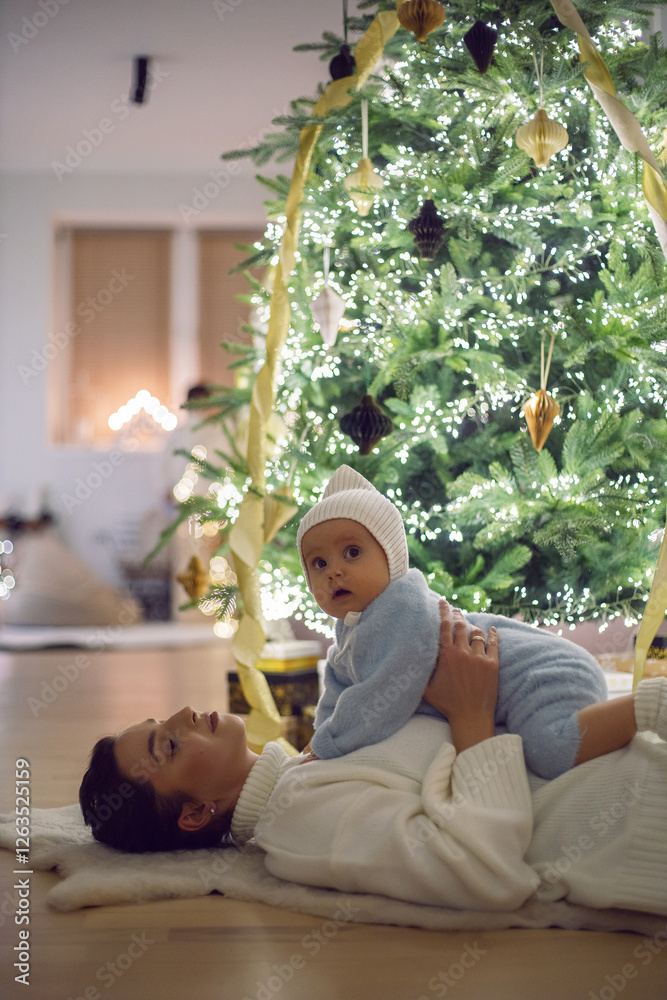 A mother in a white sweater lying with her daughter for 1 year on the floor by the Christmas tree in an apartment on New Year's Eve