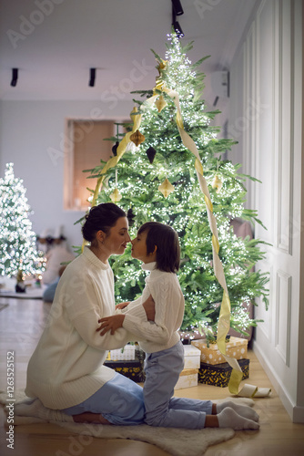 A mother in a white sweater sits with her son for 7 year on the floor by the Christmas tree in an apartment on New Year's Eve