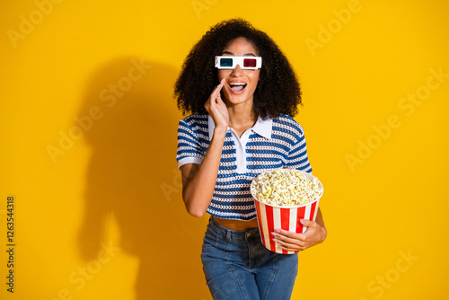 Joyful young woman in stylish casual attire wearing 3D glasses holds popcorn bucket while smiling against a bright yellow background