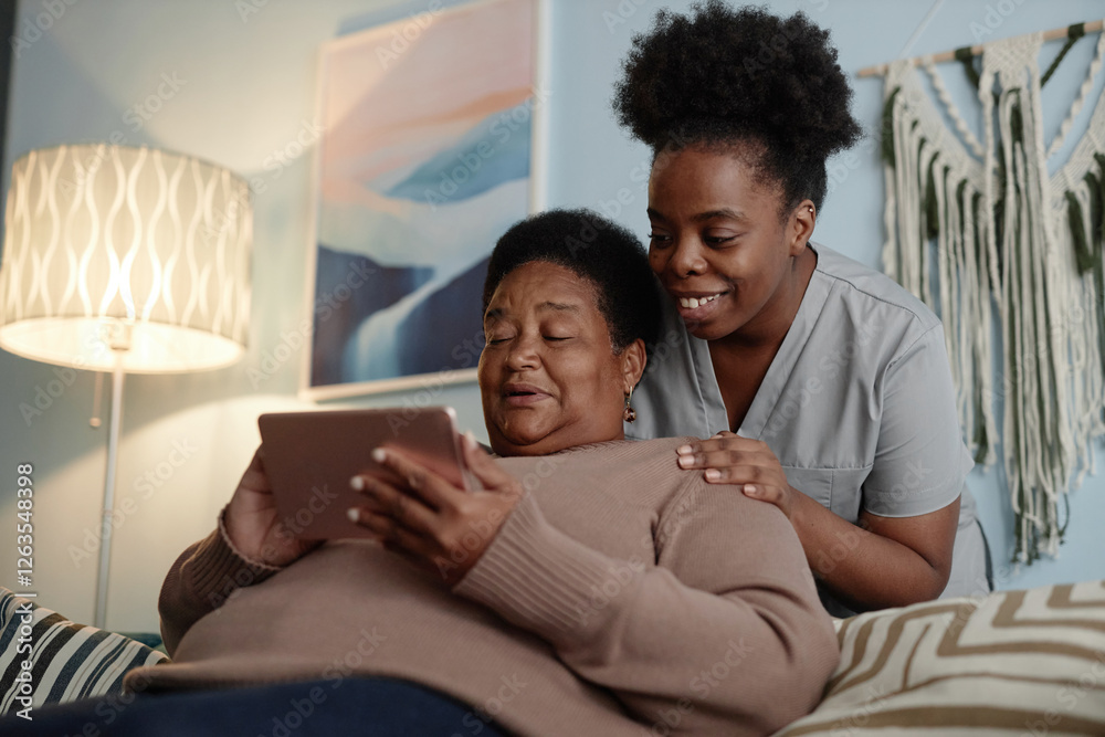 Fototapeta premium Medium shot of African American elderly woman learning how to use online payment services on tablet computer with help of supportive female social worker, while sitting on couch in cozy apartment
