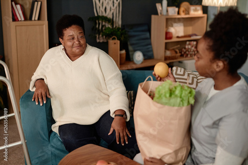 Shot in warm tones of elderly African American woman with physical disability receiving grocery delivery from accommodating female social worker at home in living room