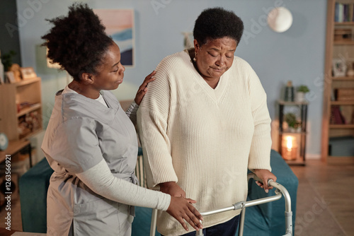 Medium shot of accommodating Black female social worker helping senior woman walking around with frame while providing home care for elderly