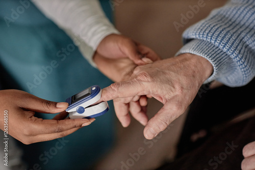Close up on wrinkled hand of elderly man placing fingertip on oximeter sensor while female social worker measuring blood oxygen level during home care visit photo