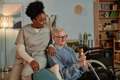 Upbeat Black female social worker helping senior male wheelchair user with phone while looking at smartphone screen sitting on couch together in living room